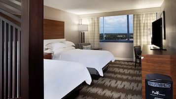 A hotel room with two beds, a desk, a chair, a TV, and a view of the city.