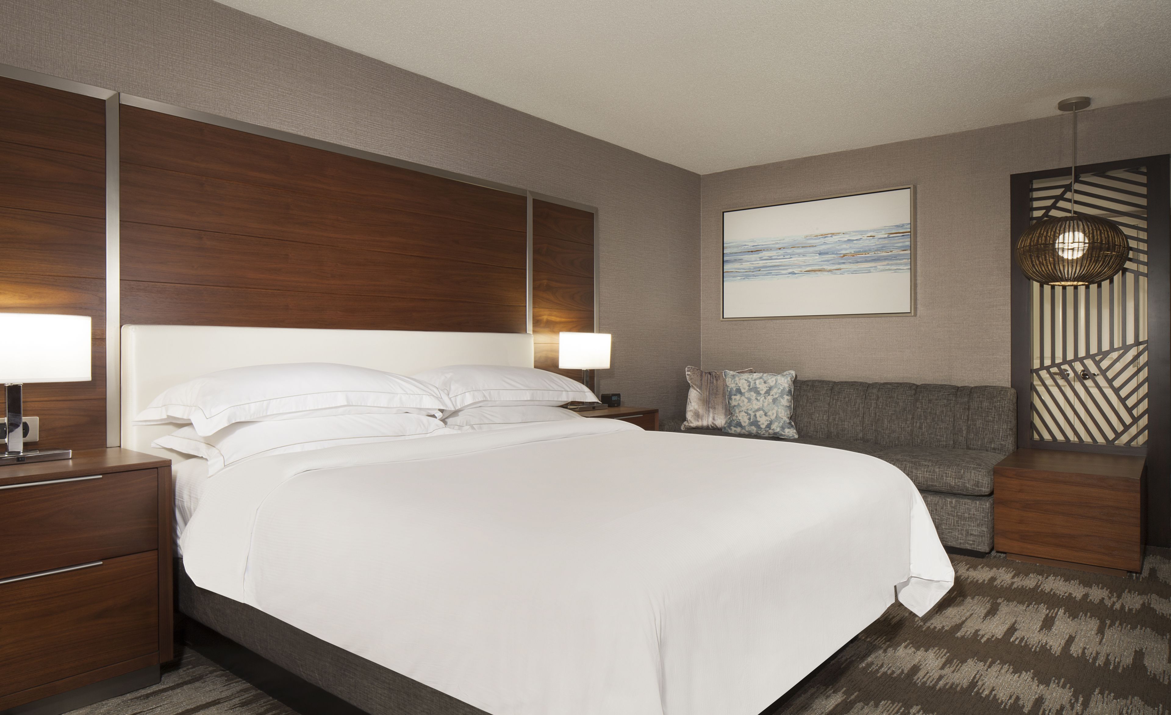 A modern hotel room with a large bed, bedside tables, a sofa, a wall-mounted artwork, and a decorative wall panel.