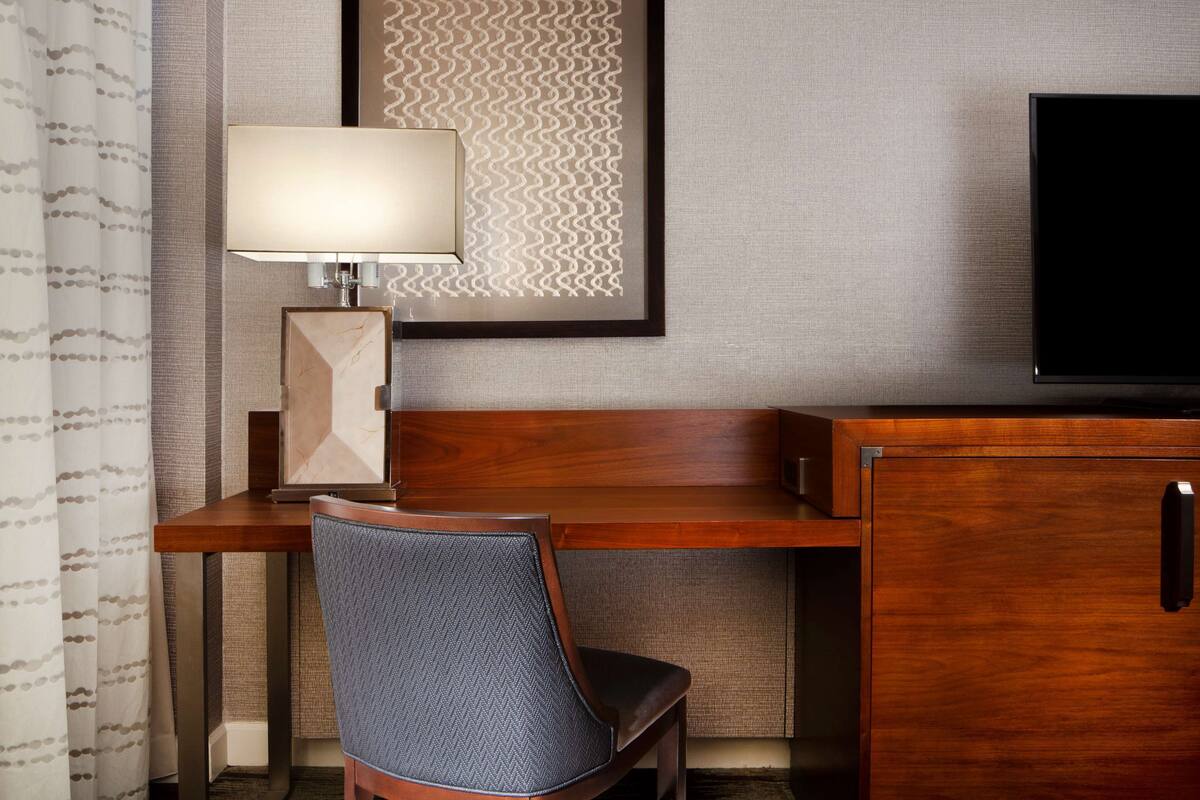 A hotel room with a wooden desk, a chair, a television, a lamp, and a framed artwork on the wall.