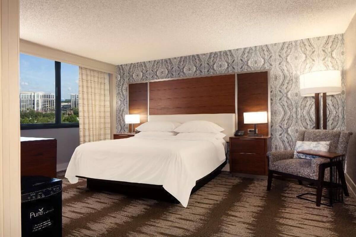 A hotel room with a large bed, two bedside tables with lamps, a chair, and a view of buildings through a window.