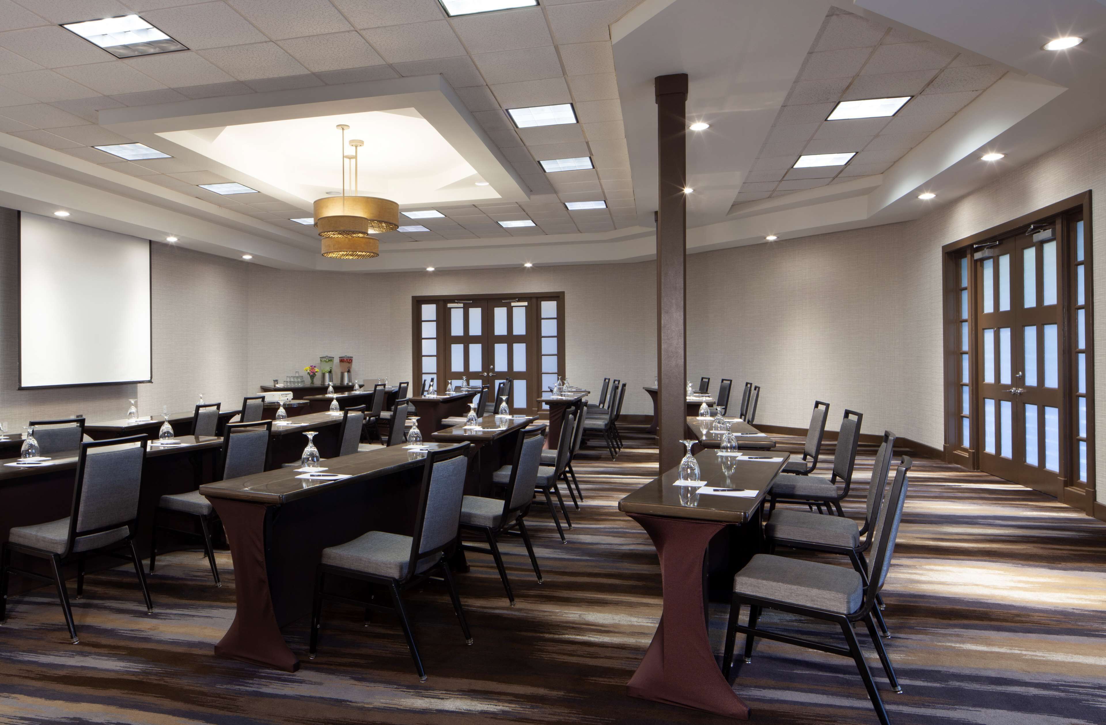 A conference room with multiple tables and chairs, a projector screen, and large windows with blinds.