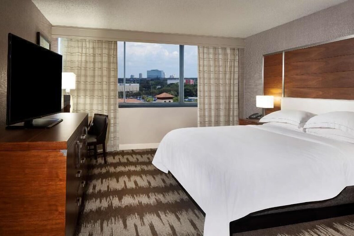 Habitación de hotel con una cama grande, televisor, escritorio y vista a la ciudad.