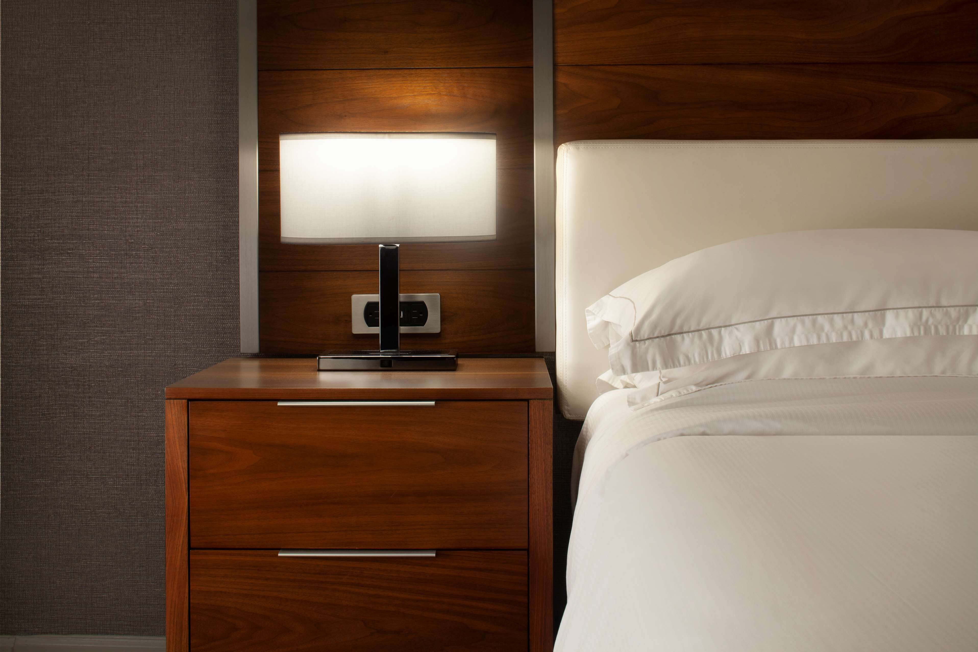 A neatly made bed with white linens, a wooden nightstand with a modern lamp, and a wall panel.