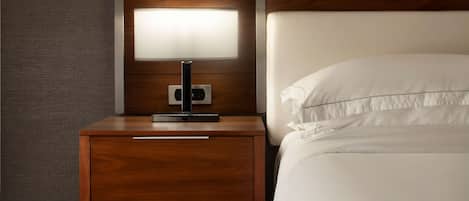 A neatly made bed with white linens, a wooden nightstand with a modern lamp, and a wall panel.