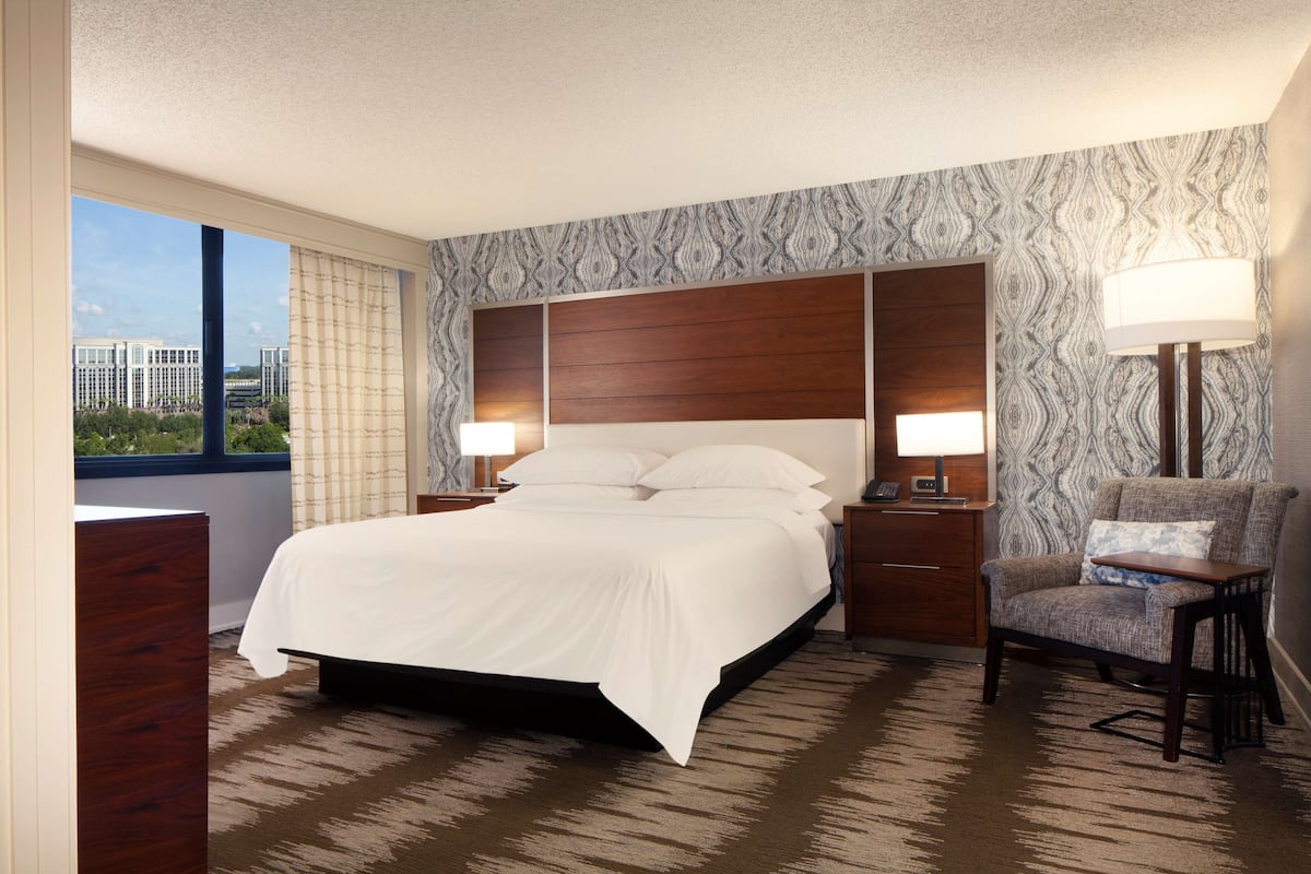 A hotel room with a large bed, two bedside tables with lamps, a chair, and a view of buildings through a window.