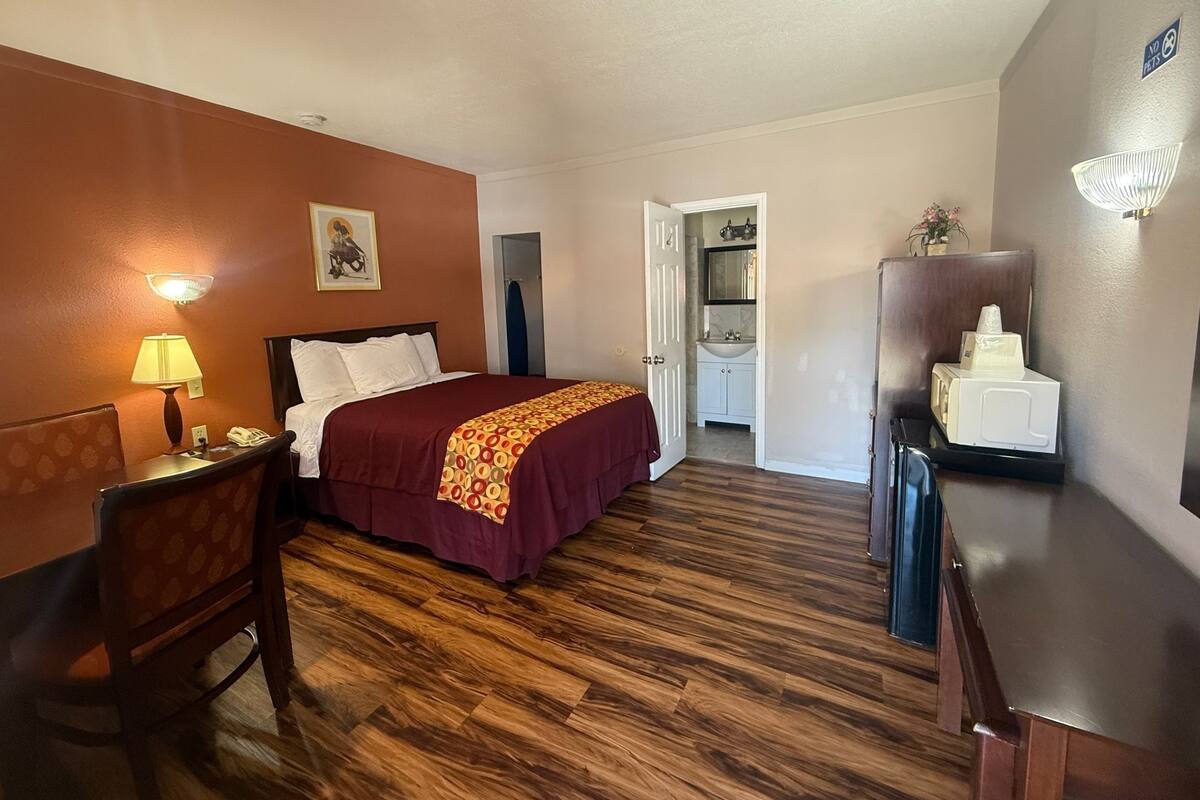 A hotel room with a bed, a desk with a lamp, a chair, a microwave, and a small refrigerator.