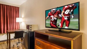 A hotel room with a flat-screen TV mounted on a wooden dresser, a desk with a chair, and a lamp. - Red Roof Inn Winchester - Lexington East (Winchester)