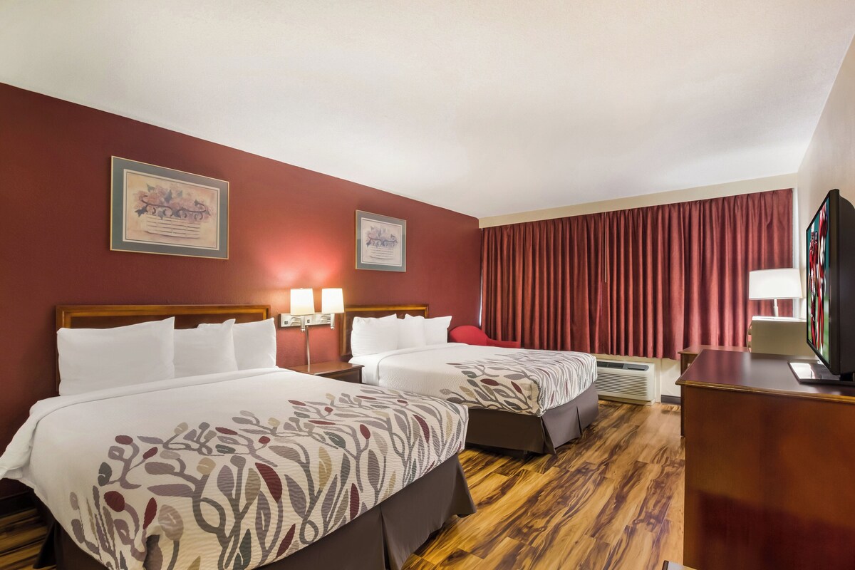 A hotel room with two beds, a TV, a desk, and a window with curtains.