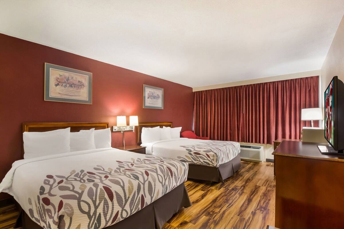 Ein Hotelzimmer mit zwei Betten, einem Fernseher, einem Schreibtisch und einem roten Vorhang.