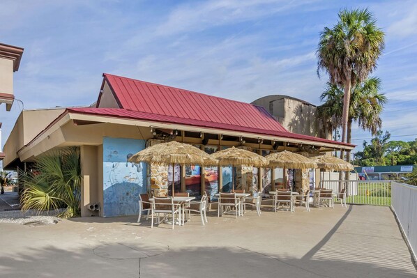 Un restaurante frente a la playa con mesas al aire libre, palmeras y techo rojo.