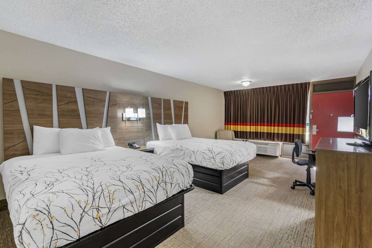 A hotel room with two beds, a desk, a chair, and a TV.