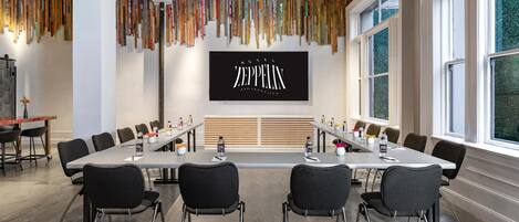 A conference room with a long table, chairs, a projector screen, and a wall with hanging batons.