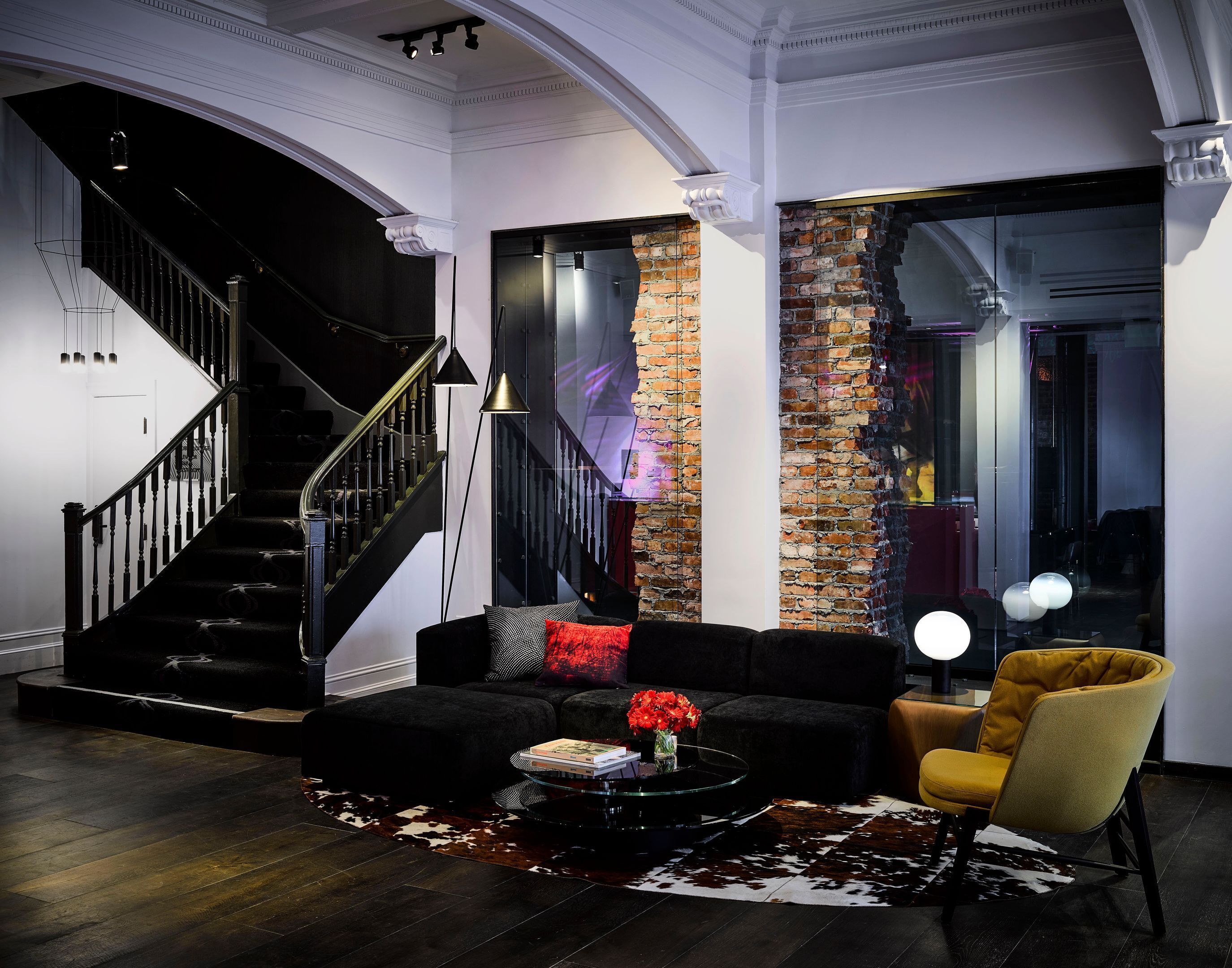 A modern living room with a black staircase, a brick accent wall, a black sofa, a yellow armchair, and a glass coffee table.