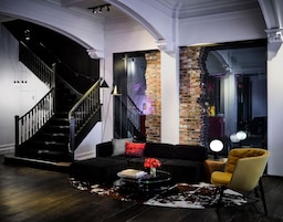A modern living room with a black staircase, a brick accent wall, a black sofa, a yellow armchair, and a glass coffee table.