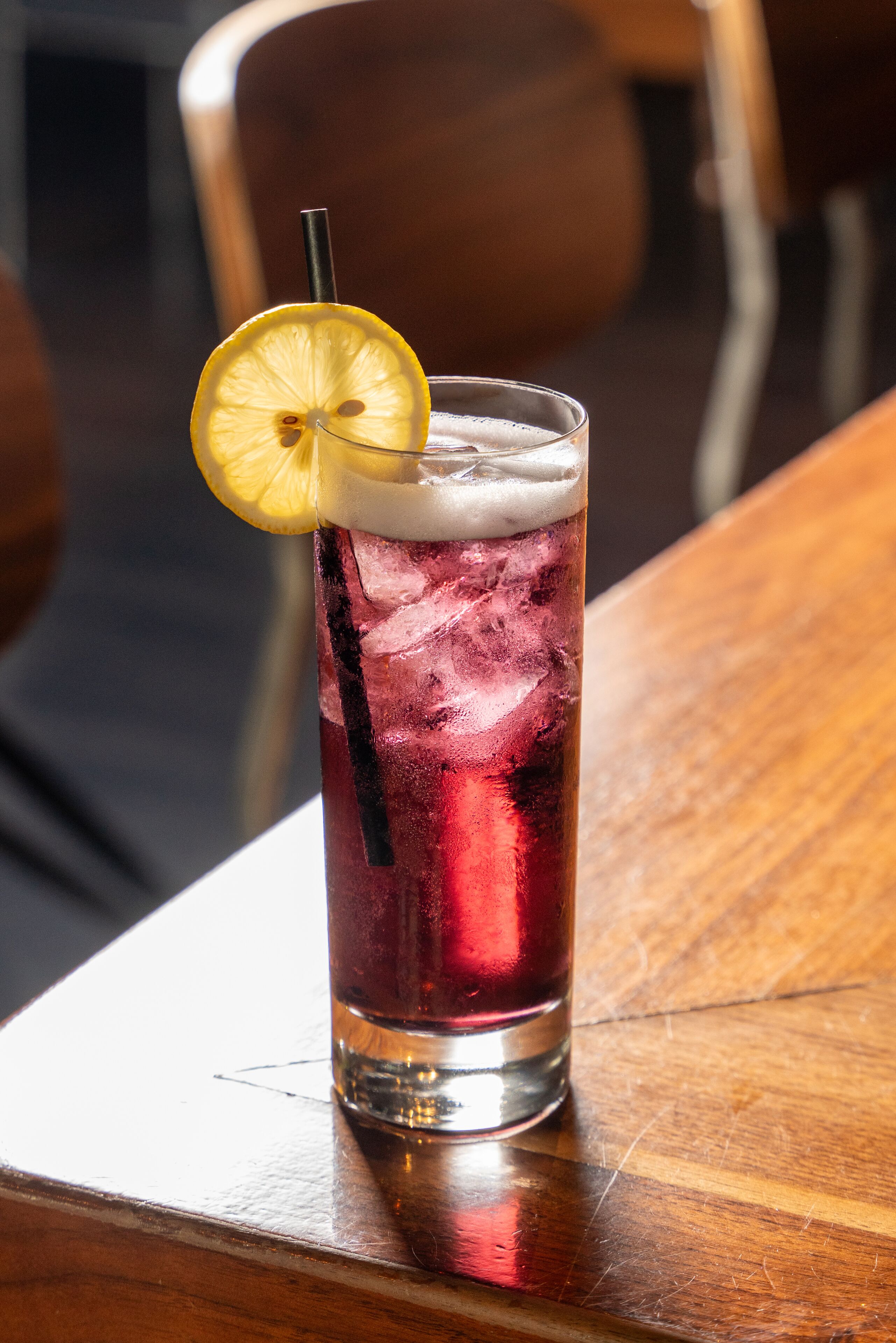 A tall glass with a dark beverage, ice cubes, and a lemon slice garnish on a wooden table.