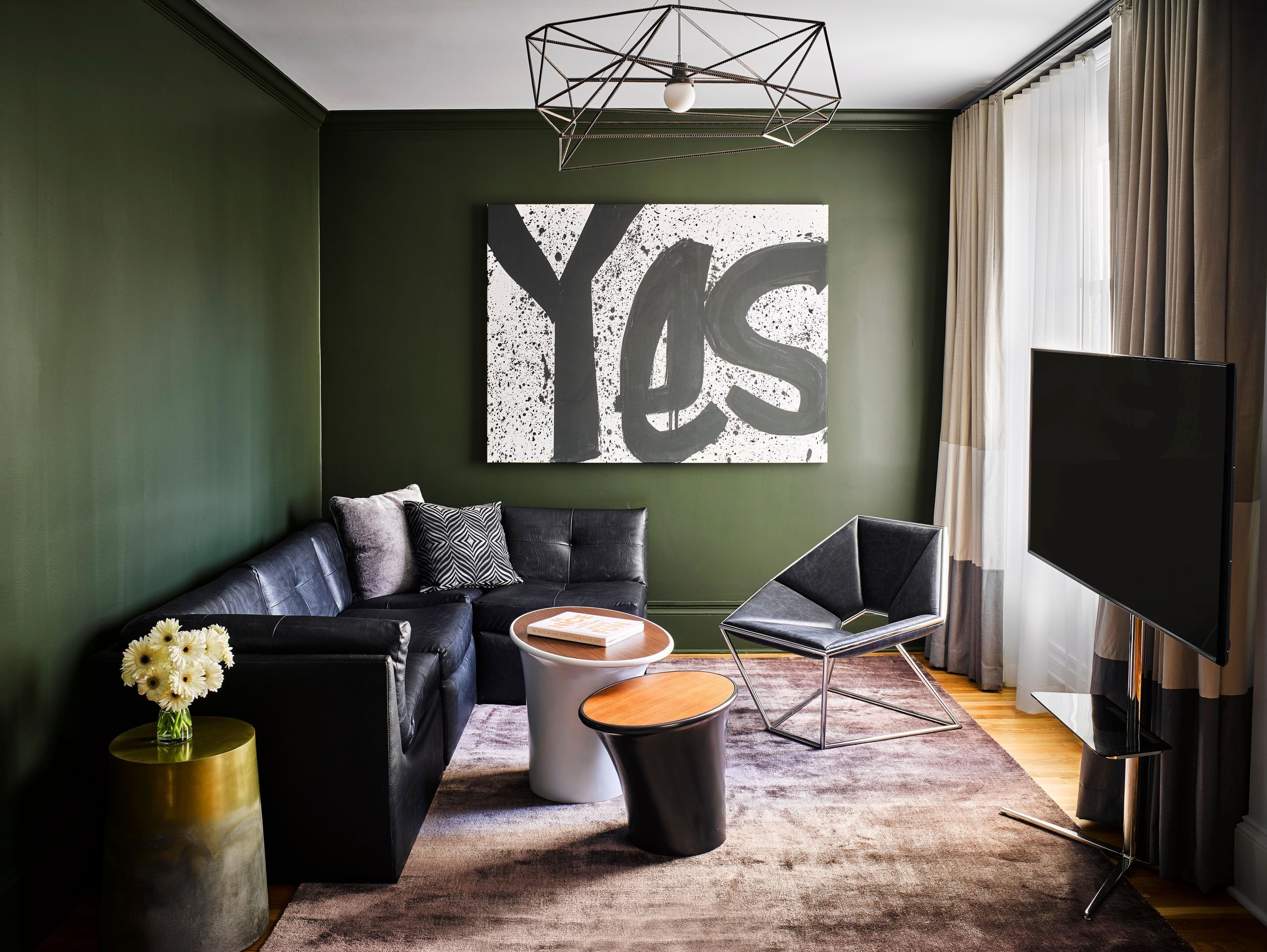 A modern living room with a black leather sofa, a coffee table, a side table with a vase of flowers, a large abstract painting, and a flat-screen TV.