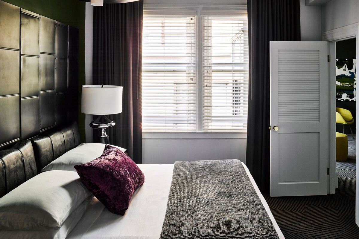 A hotel room with a bed, a lamp, a window with blinds, and a door.