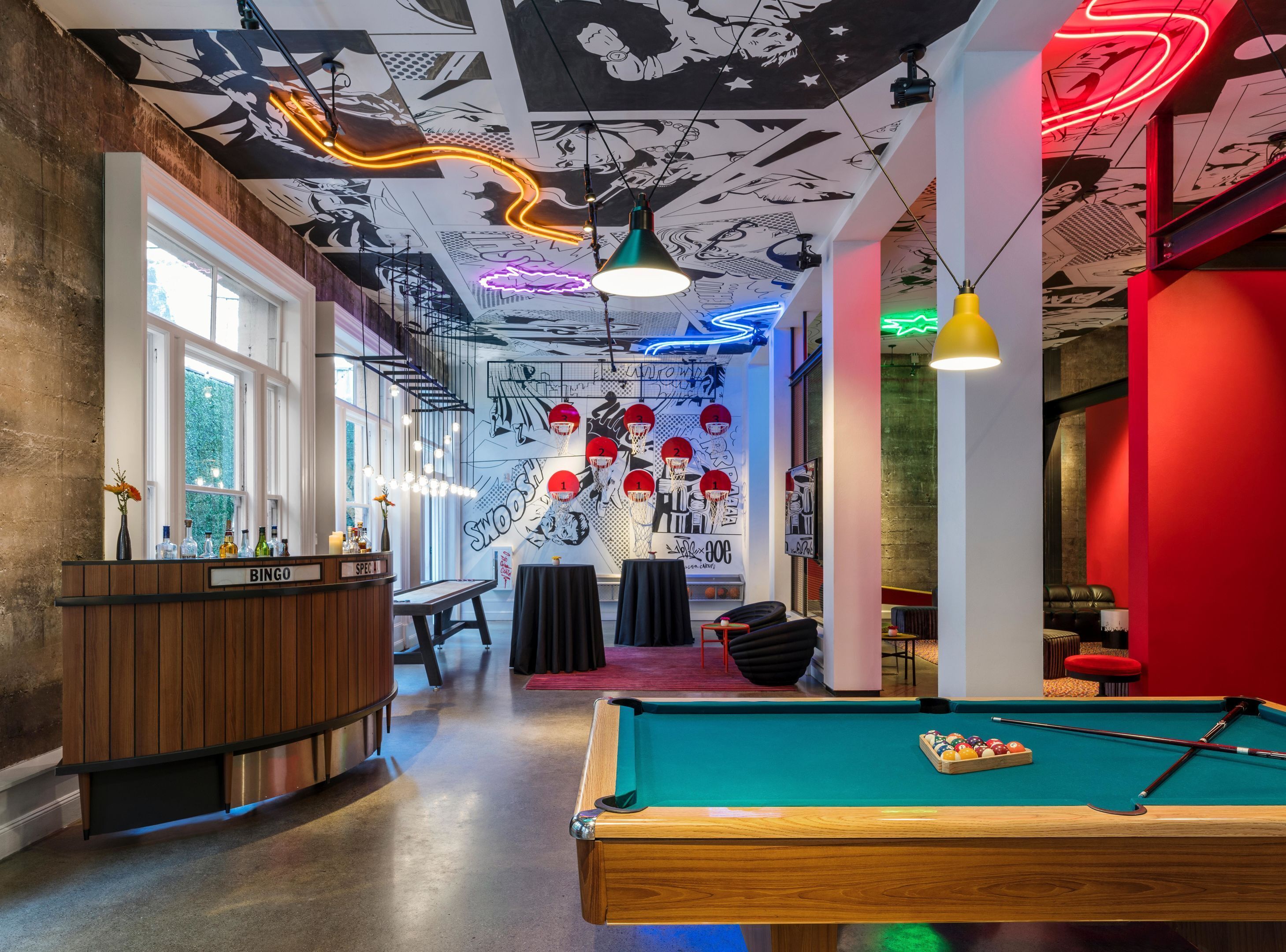 Une table de billard avec un jeu de billard et des billes, un coin bar avec une enseigne indiquant « BINGO », et une fresque murale aux motifs sur le thème du basketball.