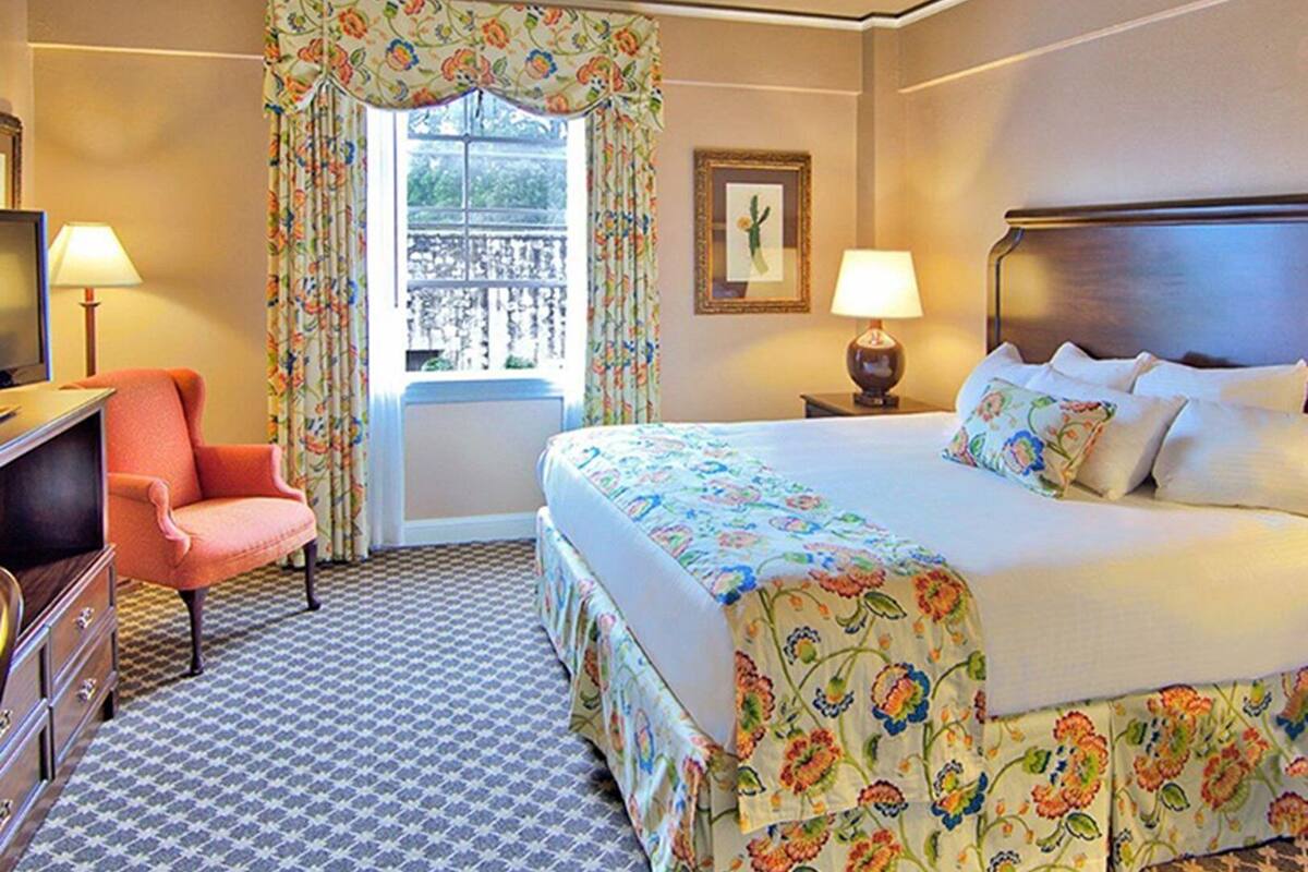 A hotel room with a bed, a desk, a chair, a window with curtains, a nightstand, and a lamp.