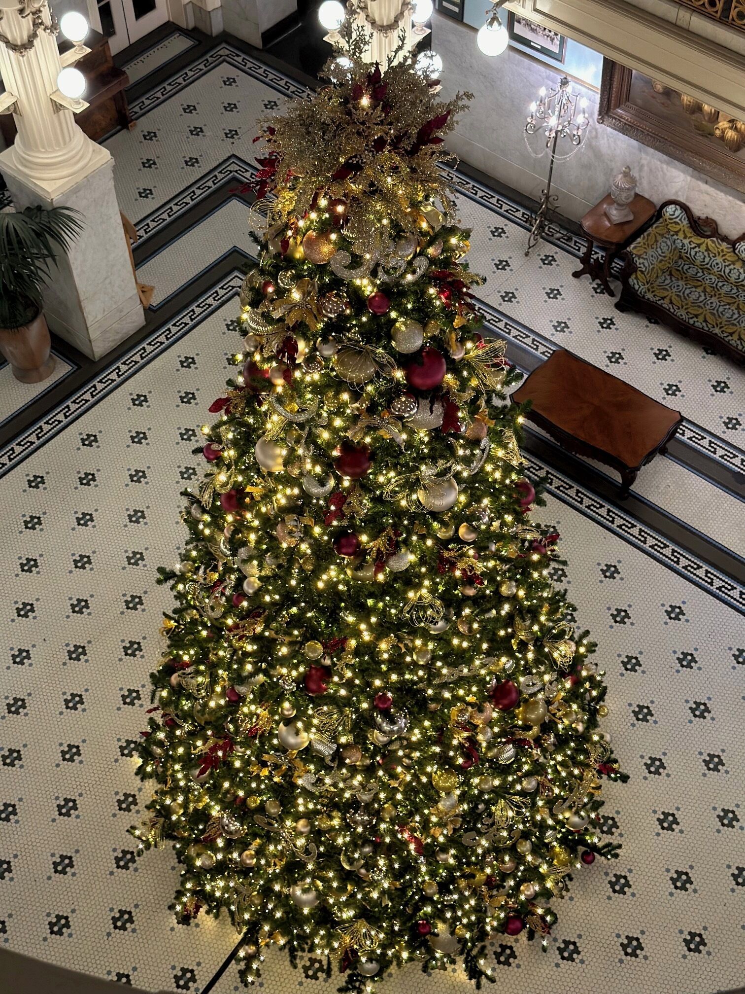 A large, ornately decorated Christmas tree with lights and ornaments, situated in a spacious lobby with patterned flooring and classical architectural details.