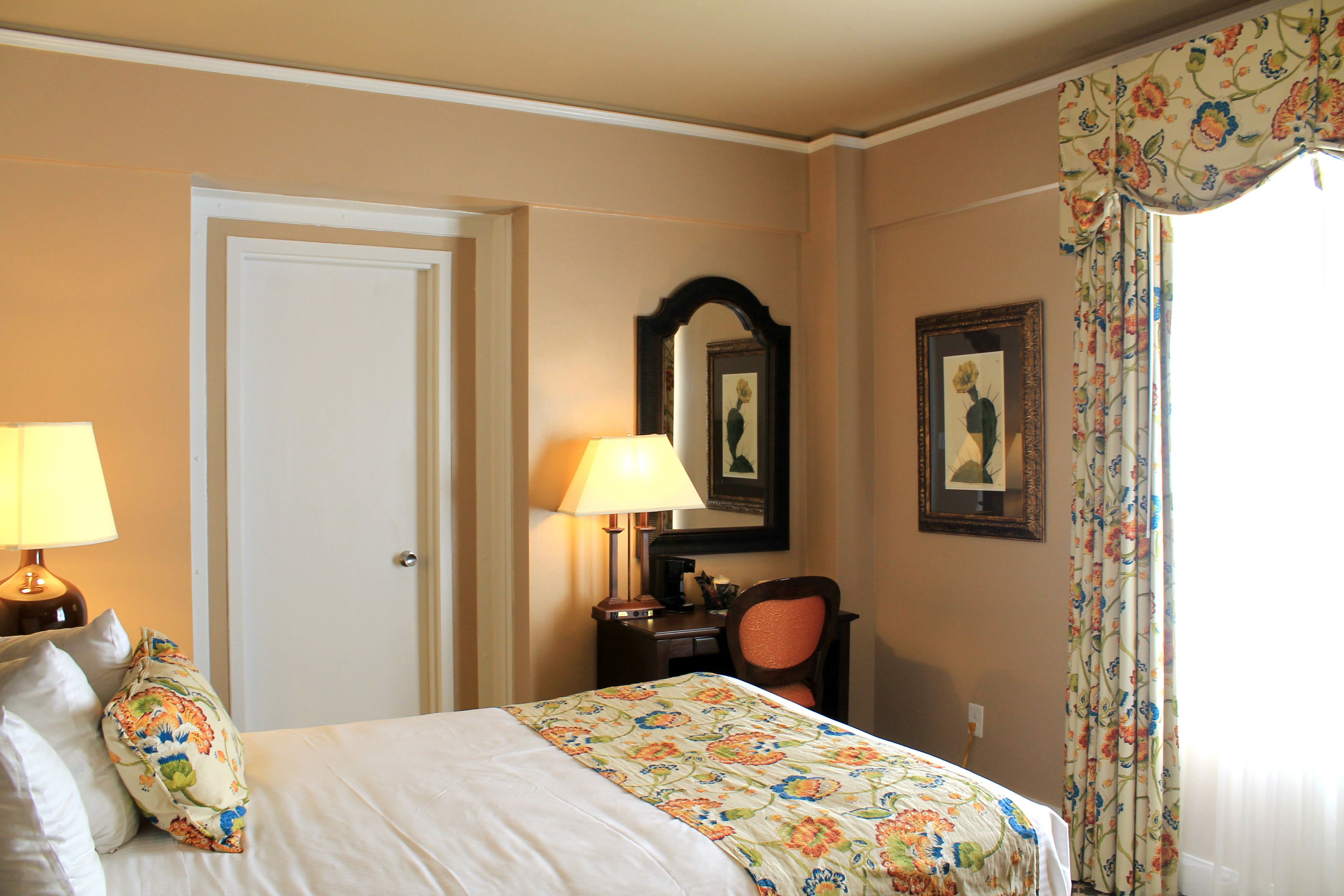A hotel room with a bed, two lamps, a desk, a chair, a door, and a window with curtains.