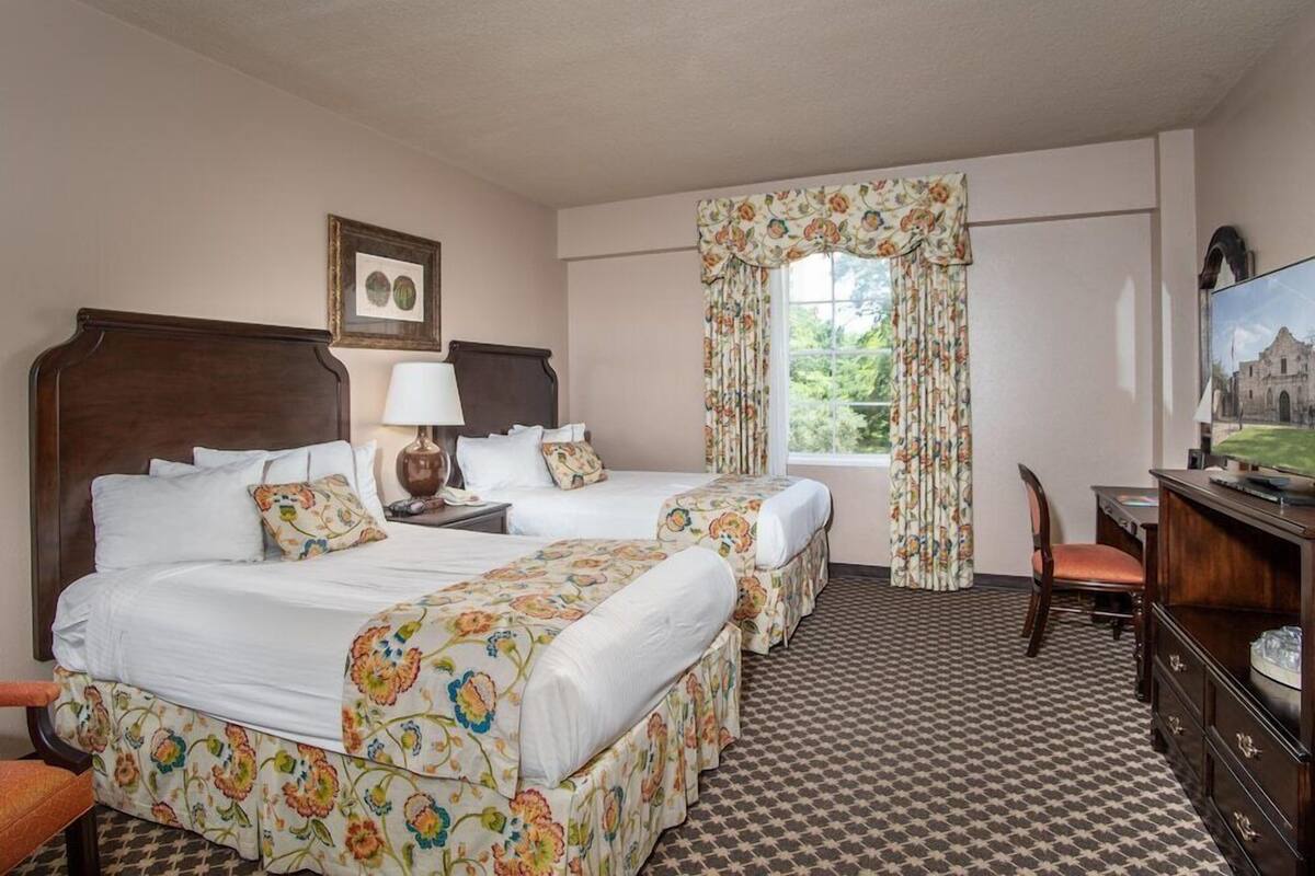 A hotel room with two beds, a desk, a chair, a television, and a window with floral curtains.