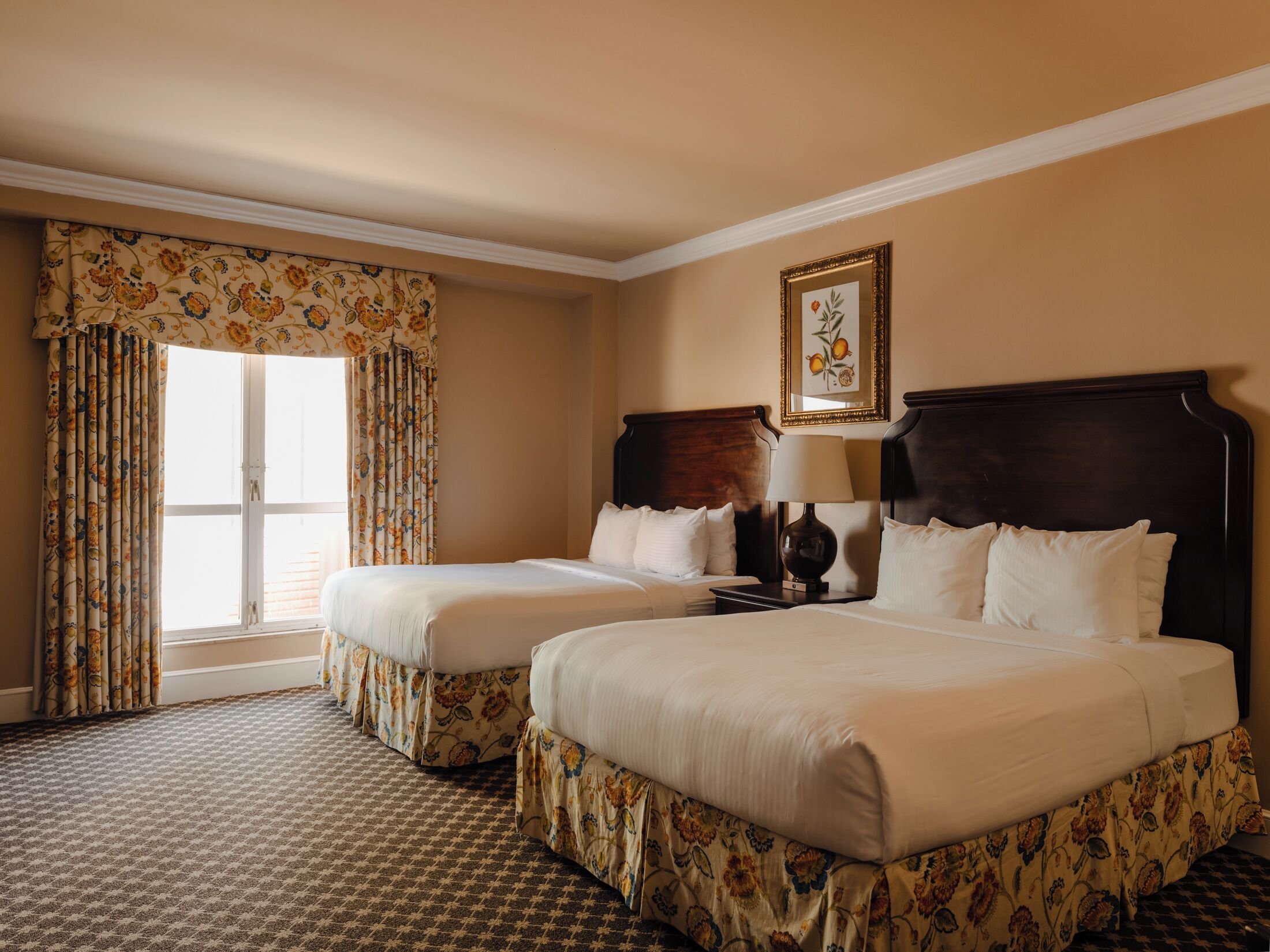 Une chambre d’hôtel avec deux lits, une grande fenêtre avec des rideaux, un tableau encadré au mur et une moquette à motifs.