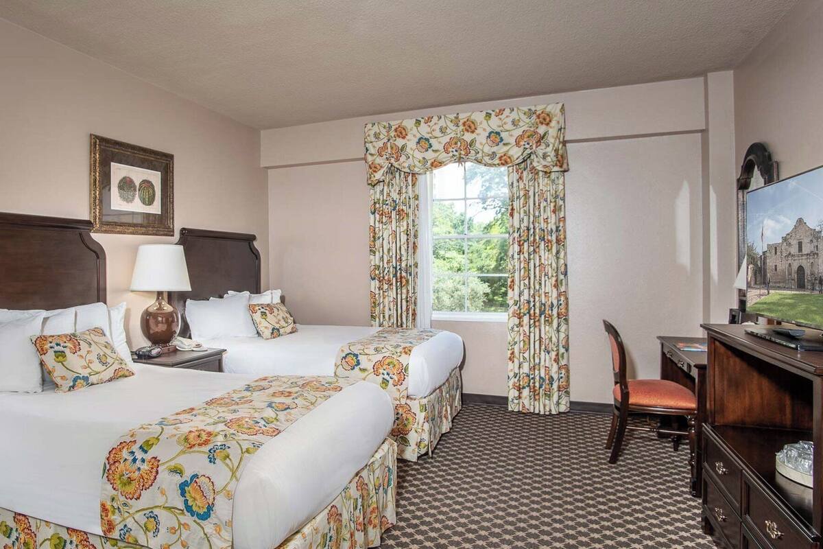 A hotel room with two beds, a desk, a chair, a television, and a window with floral curtains.
