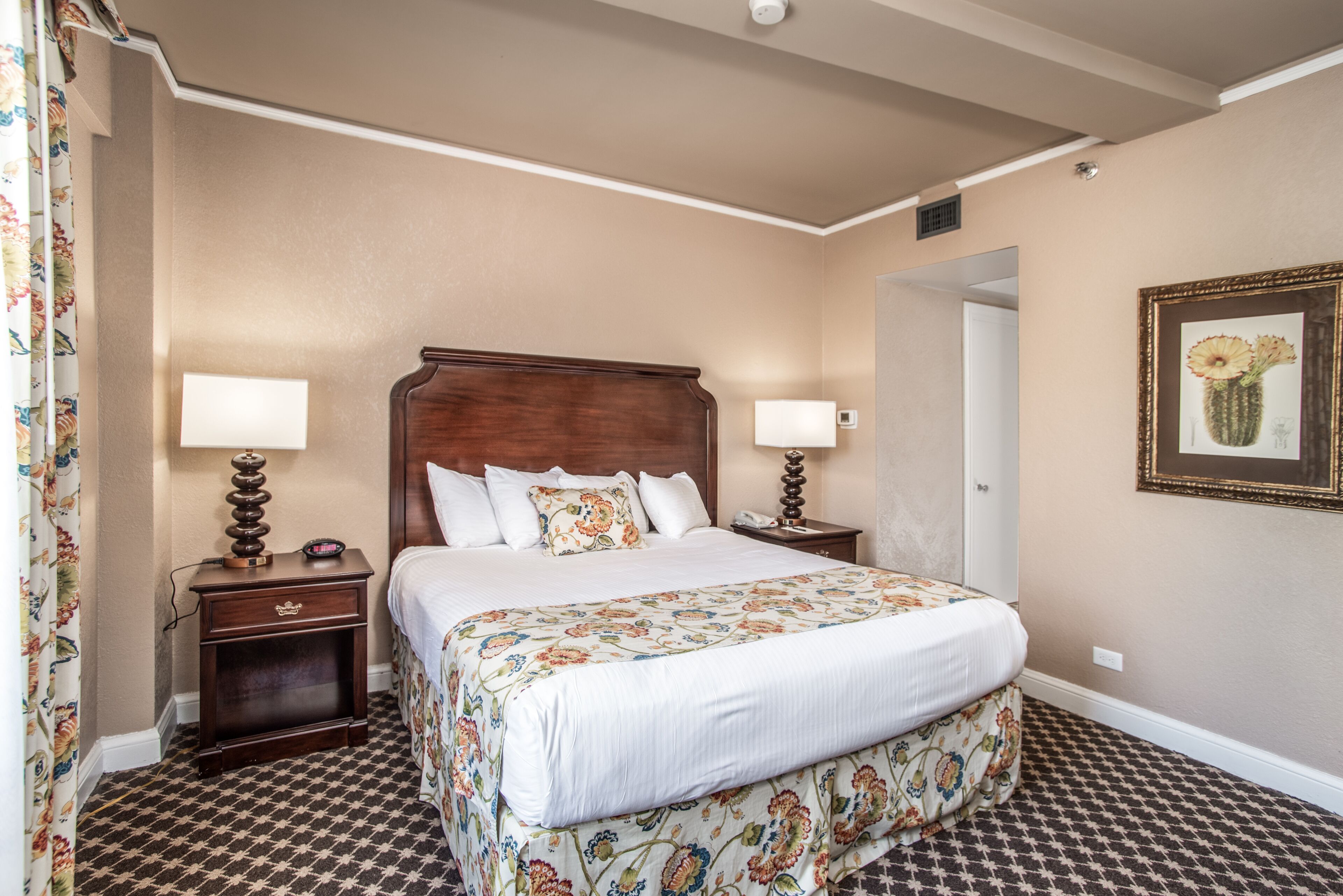 A hotel room with a large bed, two bedside tables, a nightstand, a floral-patterned bedspread, and a framed cactus artwork on the wall.