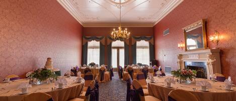 A banquet hall with round tables, chairs with sashes, a chandelier, and a fireplace.