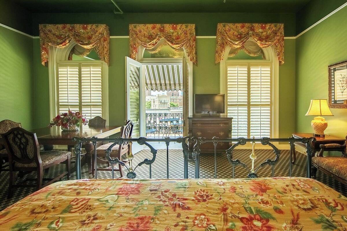 A hotel room with a large bed, two windows with curtains, a balcony, a TV, and a lamp.