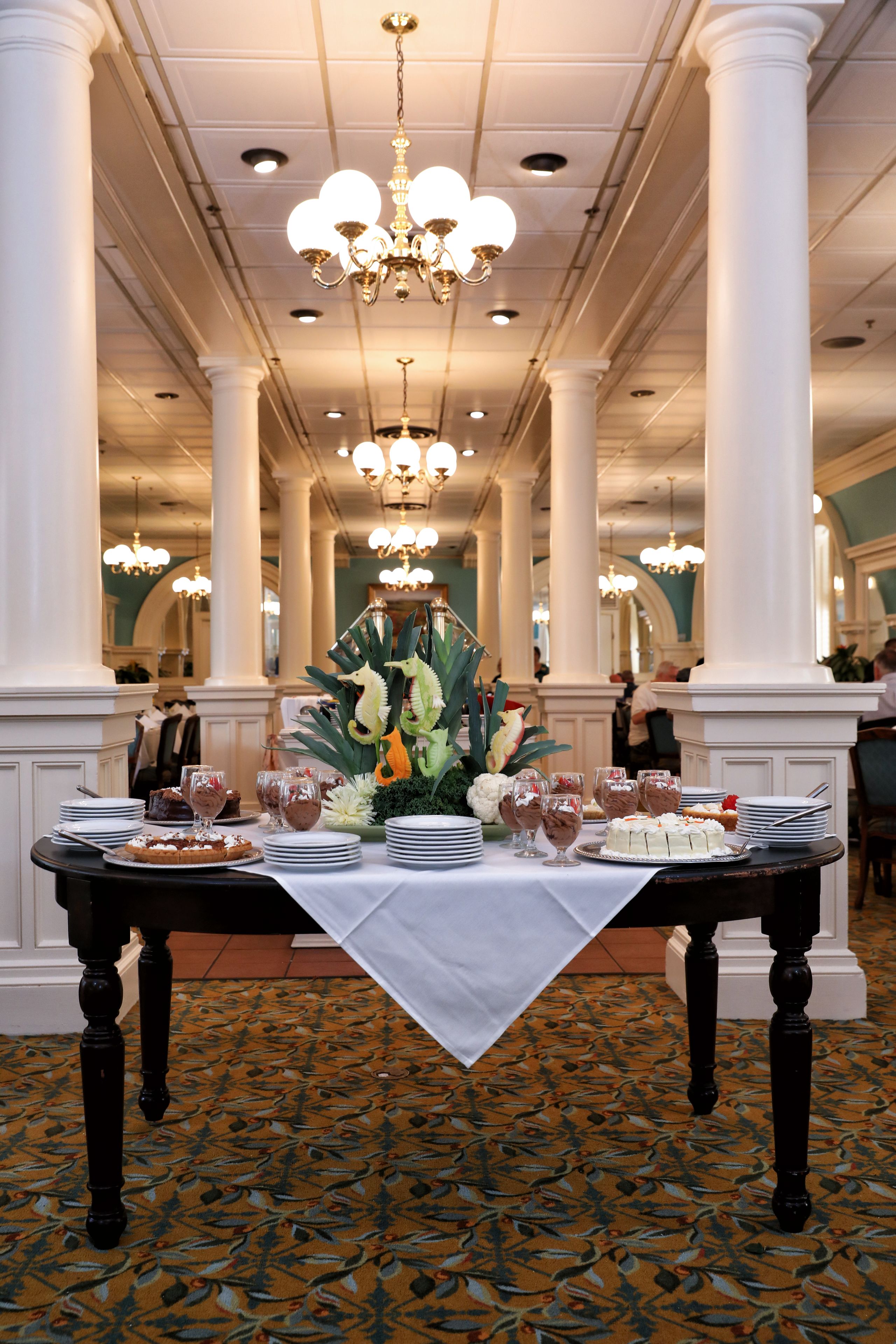 A formal dining setup with a table set for multiple guests, surrounded by white columns and chandeliers.