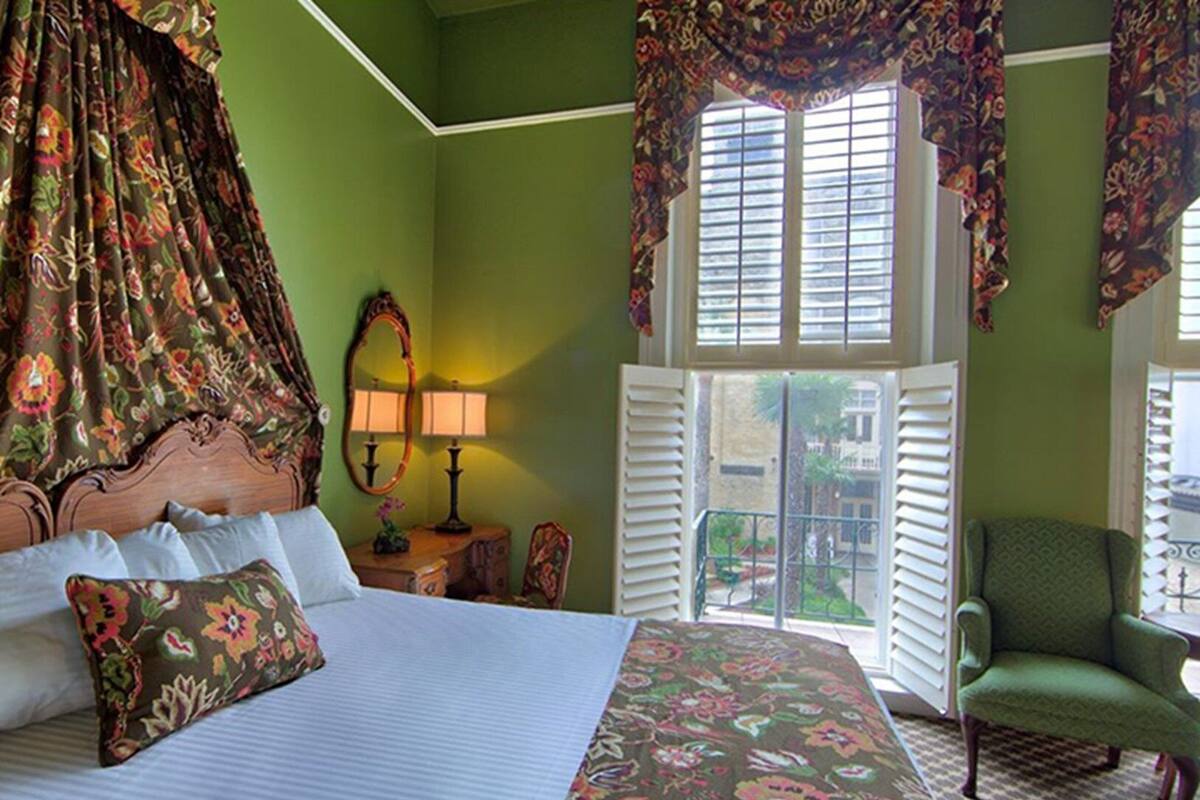 A bedroom with a large bed, floral curtains, a window with shutters, a chair, and a mirror.