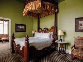 Una cama con dosel y cortinas, una mesita de noche con lámpara, una silla e imagen enmarcada en la pared.