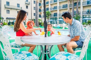Una familia cenando en una mesa al aire libre con vista a la piscina, rodeada de sillas blancas con diseños azules.