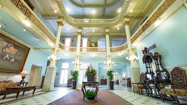 A grand lobby with a central staircase, ornate columns, and a chandelier. There are potted plants and a large painting on the walls.