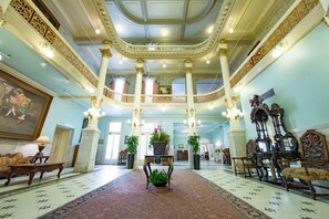 Un'imponente hall con una scalinata centrale, colonne decorate e un lampadario. Si notano piante in vaso e un grande quadro appeso alle pareti.