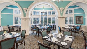A dining area with multiple tables and chairs, large windows, and a chandelier.