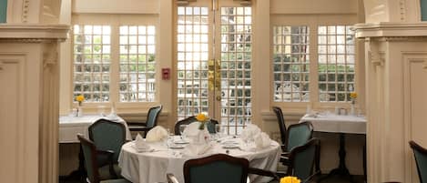 Un comedor con mesas dispuestas para cuatro comensales, cada una con mantel blanco, cubertería y cristalería. Las sillas tienen tapicería verde y un candelabro cuelga del techo.
