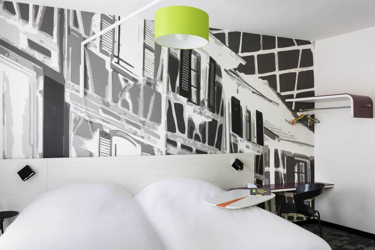 Hotelkamer met een bed, bureau, stoel, lamp en een muur met een zwart-wit abstract ontwerp.