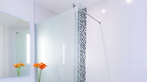 Un baño moderno con ducha de mampara de vidrio, un lavabo blanco con espejo y un jarrón con flores naranjas.