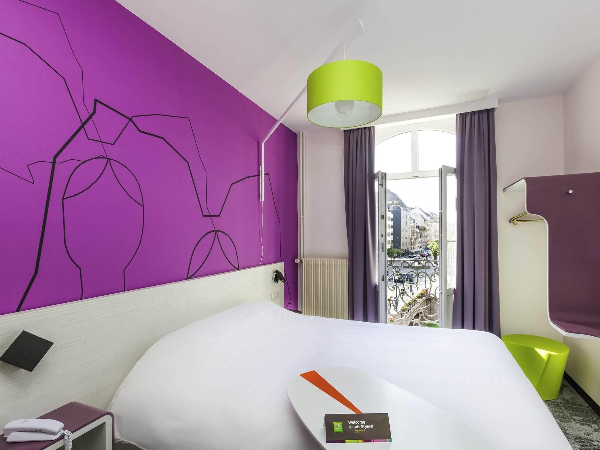 Una habitación de hotel con una cama, una mesita de noche con un teléfono, una pared con una obra de arte abstracta, una ventana con cortinas y vistas a edificios.