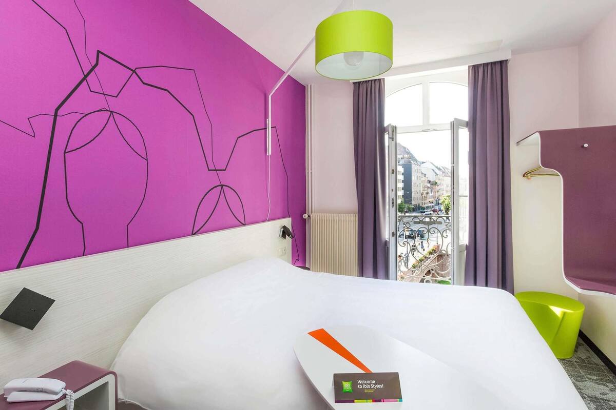Hotelkamer met een bed, nachtkastje met telefoon, felgroene lamp, raam met gordijnen en uitzicht op gebouwen.