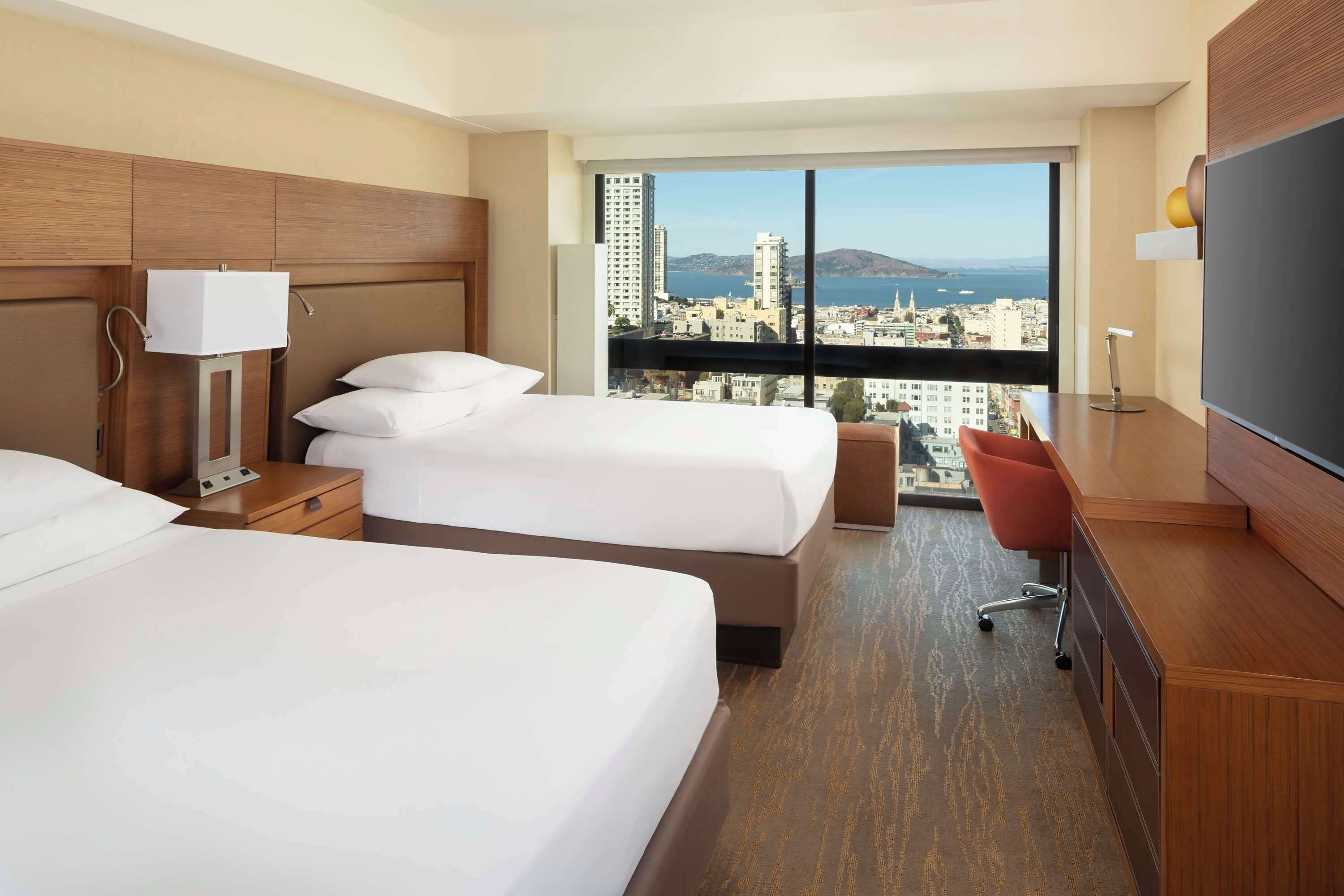 A hotel room with two beds, a desk, a chair, a TV, and a large window with a city view.