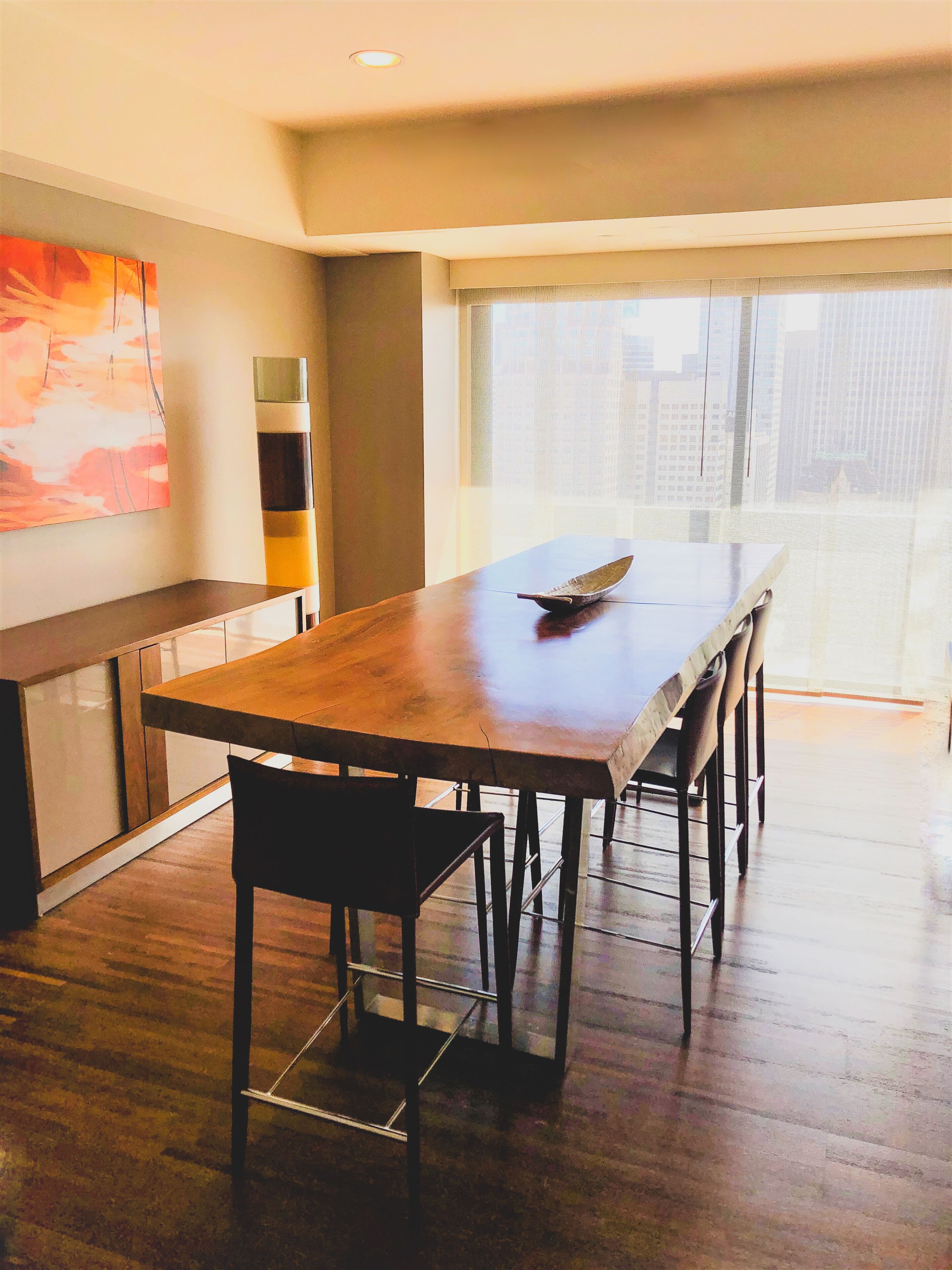 A modern dining area with a wooden table and chairs, a large window with a city view, and a contemporary art piece on the wall.