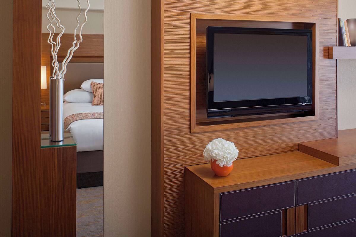 A modern hotel room with a large flat-screen TV mounted on a wooden wall, a small orange vase with white flowers on a wooden console, and a bed with white linens and a patterned pillow.