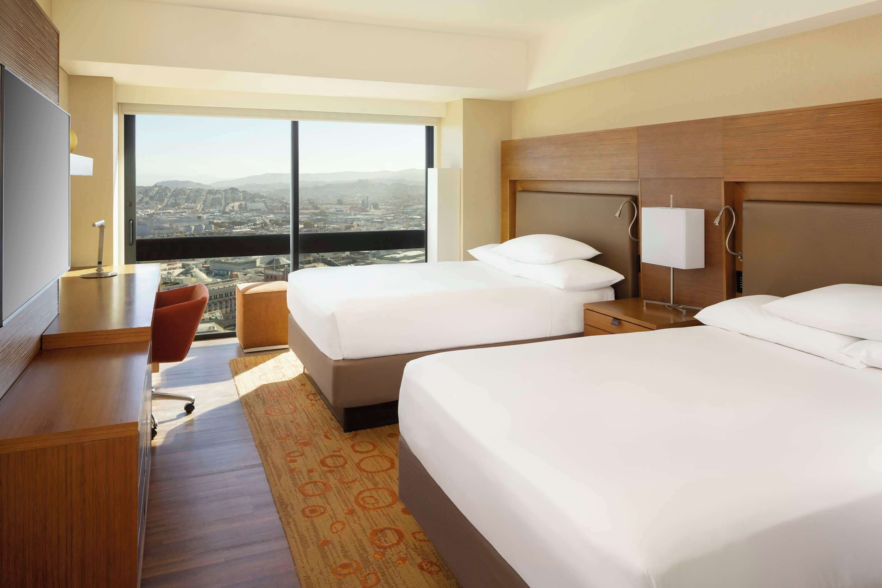 Ein Hotelzimmer mit zwei Betten, einem Schreibtisch, einem Fernseher und einem großen Fenster mit Blick auf die Stadt.
