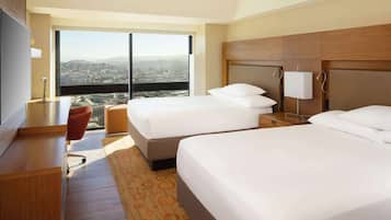 Ein Hotelzimmer mit zwei Betten, einem Schreibtisch, einem Fernseher und einem großen Fenster mit Blick auf die Stadt.