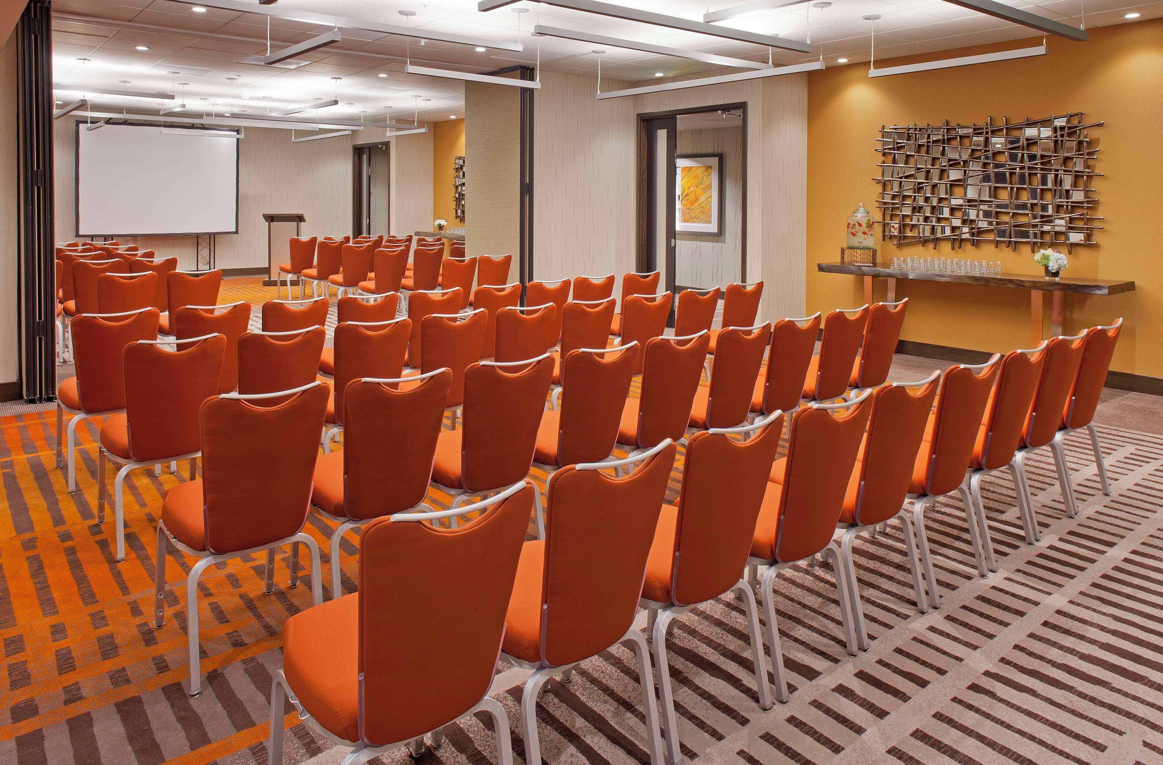 A conference room with rows of orange chairs, a white projection screen, and a wooden wall display.