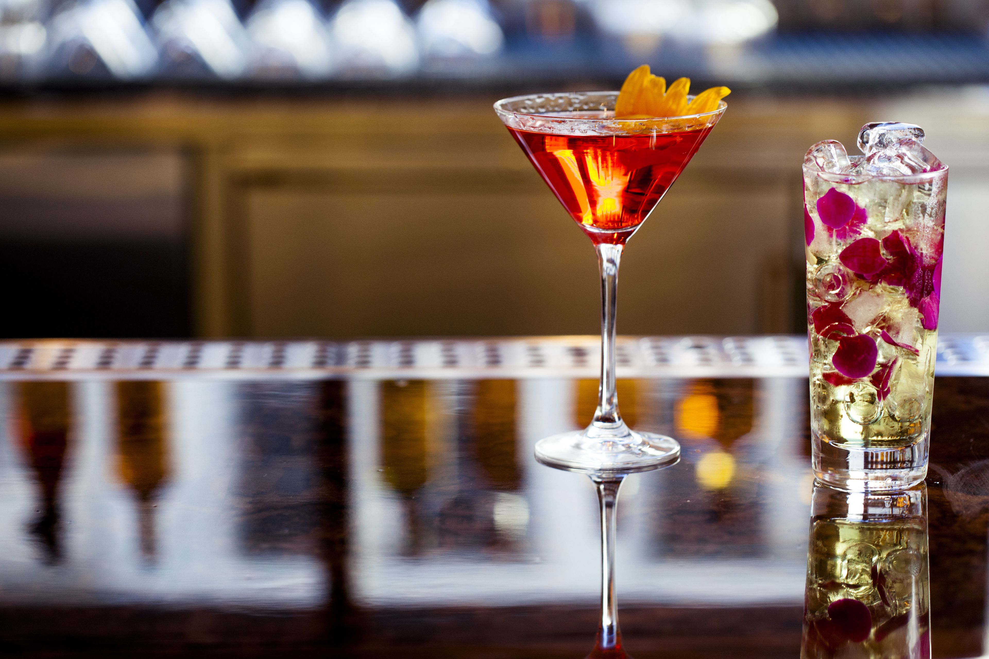 Dos bebidas sobre una barra: una en una copa de martini y otra en un vaso alto con hielo y pétalos de flores.